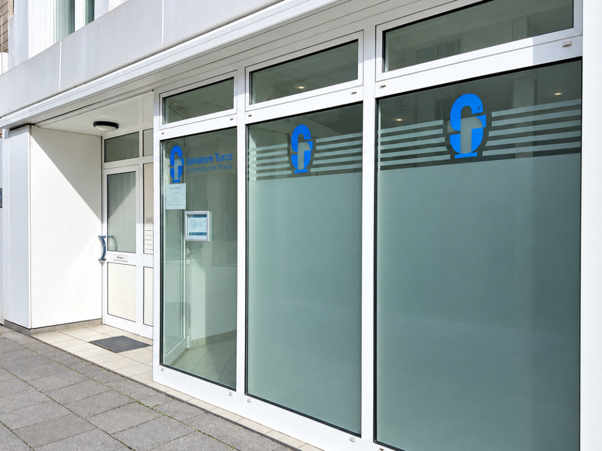 Modernes Arzt- oder Bürogebäude-Eingang mit großflächigen Glasscheiben und weißer Fassade und Firmenlogo.