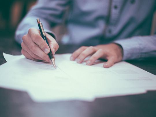 Eine Person schreibt mit einem Stift auf Papier. Sie trägt ein Hemd und konzentriert sich auf das Dokument.