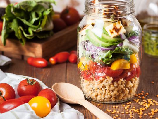 Ein bunter Salat im Glas, geschichtet mit Gemüse, Getreide und Käse. Daneben liegen frische Tomaten und ein Holzlöffel.