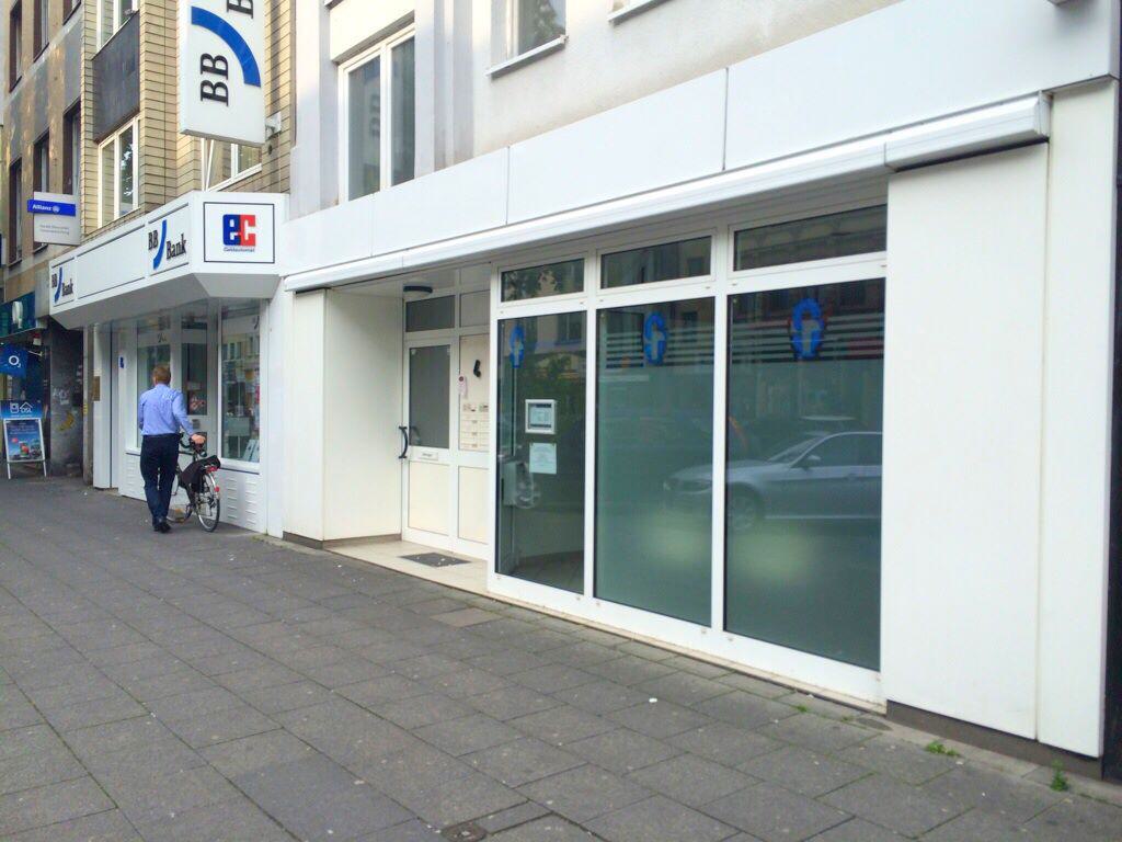 Die Fassade eines Gebäudes mit einer großen Fensterfront und einer Eingangstür. Die Fenster sind teilweise undurchsichtig, und es gibt zwei blaue Logos auf den Fenstern.