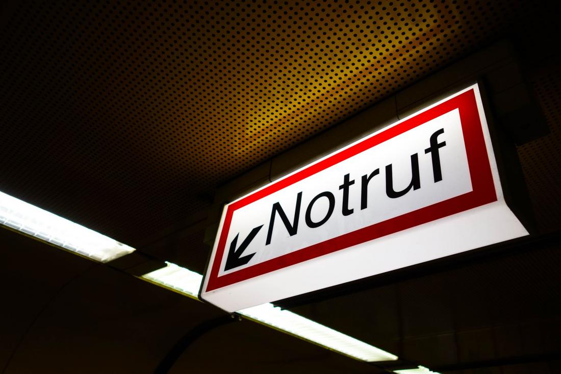Ein beleuchtetes Schild mit der Aufschrift "Notruf" und einem Pfeil nach links. Das Schild befindet sich in einem Gebäude mit einer perforierten Decke und Leuchtstofflampen.